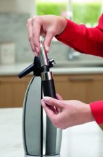 פעולת מילוי בלון הגז (CO2) לתוך סיפון הסודה. רואים יד לובשת חולצה אדומה ושעון יד אלגנטי עם רצועת מתכת, העוסקת בהרכבת המכשיר במטבח מודרני.