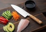 מבט תקריב על סכין Sico Santoku מוכנה לעבודה, ליד ירקות חתוכים לסושי ונתחי דג גולמיים על משטח עץ.