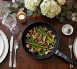 שולחן אוכל יוקרתי ערוך עם כלים לבנים, נרות ופרחים, שבמרכזו מונחת מחבת סוויס דיאמונד עם אספרגוס ופטריות צלויים.