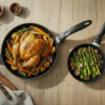 ארוחה משפחתית: מחבת 24 ס"מ עם פטריות ואספרגוס לצד מחבת 28 ס"מ עם עוף צלוי שלם וירקות שורש על שולחן עץ.