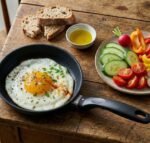 ארוחת בוקר כפרית: ביצת עין מתובלת בתוך מחבת קטנה Swiss Diamond 18 ס"מ, מוגשת על שולחן עץ לצד ירקות ולחם.